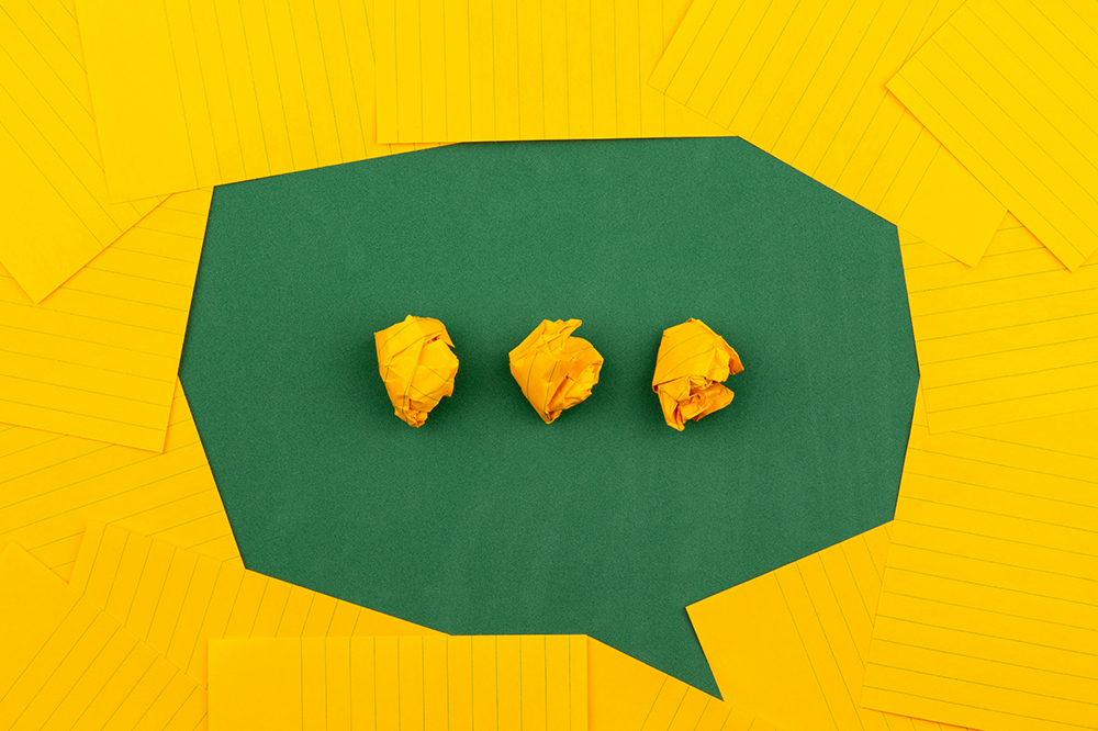 Yellow sheets of lined paper arranged on a green surface so that the green are looks like a speech bubble. Three crumpled balls of paper in the middle of the speech bubble